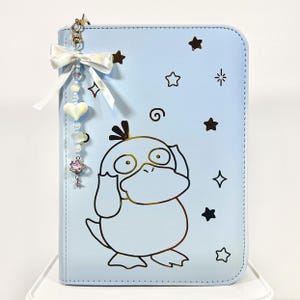 Può includere: Una copertina per notebook azzurra con un design a cartone animato di un'anatra con dettagli dorati e motivi a stella. Include un fiocco bianco e un ciondolo con perline e un cuore. La copertina è di un colore pastello.