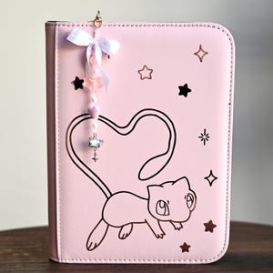 Puede incluir: Una carpeta de color rosa claro con una ilustración de líneas negras de una criatura felina cuya larga cola forma un corazón. La cubierta presenta recortes en forma de estrella y un colgante con un lazo de cinta blanca y cuentas.