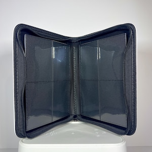 May include: Open black card binder with clear plastic sleeves. The binder is zippered and has a white border. The interior features multiple pockets for storing cards. The binder is open, displaying the interior card slots.