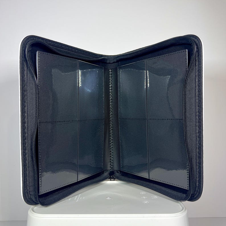 May include: Open black card binder with clear plastic sleeves. The binder is zippered and has a white border. The interior features multiple pockets for storing cards. The binder is open, displaying the interior card slots.