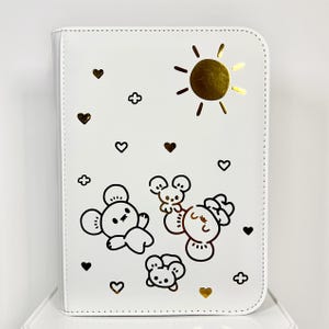 May include: White zippered binder with a gold sun, hearts, and cartoon animal illustrations. The cover features a gold sun, hearts, and four cartoon animal illustrations in black. The binder is designed for organizing papers or other items.