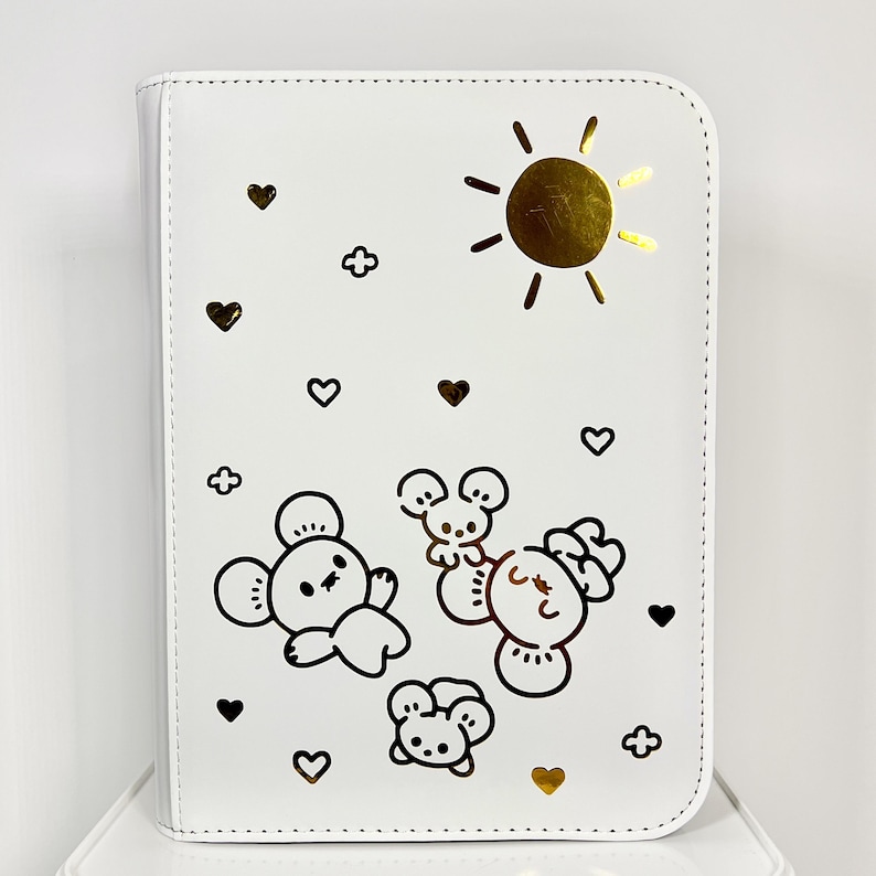 May include: White zippered binder with a gold sun, hearts, and cartoon animal illustrations. The cover features a gold sun, hearts, and four cartoon animal illustrations in black. The binder is designed for organizing papers or other items.