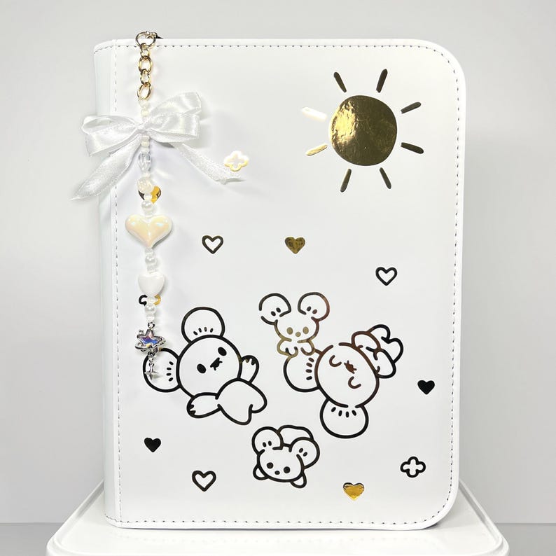 May include: White faux leather planner with a gold sun and cartoon bear illustrations. A charm with a white bow, beads, and heart-shaped accents hangs from the top. Black hearts and gold hearts are scattered across the planner's surface.