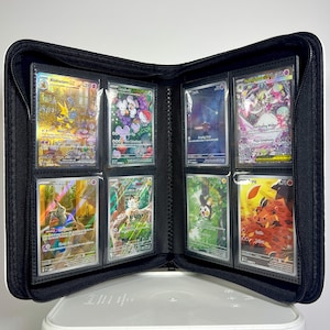 May include: Black zippered binder open to display trading cards. The cards are in clear sleeves and feature colorful illustrations of various characters. The binder is on a white surface.