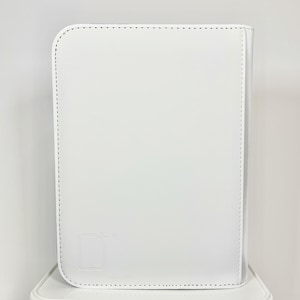 May include: A white, rectangular, leather-bound portfolio with rounded corners. The cover features a stitched border and a subtle embossed design in the lower left corner. The portfolio is sitting on a white surface.