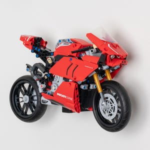 May include: A red Lego Ducati Corse motorcycle model with black wheels and a clear windscreen. The motorcycle has a detailed design with visible gears and other mechanical parts.