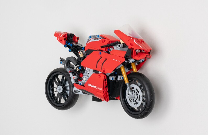 May include: A red Lego Ducati Corse motorcycle model with black wheels and a clear windscreen. The motorcycle has a detailed design with visible gears and other mechanical parts.