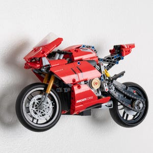 May include: A red and black LEGO Ducati Corse motorcycle model with a clear windscreen. The motorcycle has a detailed engine and working gears. The model is mounted on a white wall.