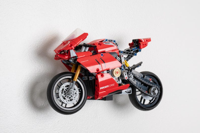 May include: A red and black LEGO Ducati Corse motorcycle model with a clear windscreen. The motorcycle has a detailed engine and working gears. The model is mounted on a white wall.