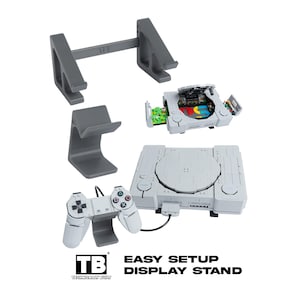 May include: A set of dark grey plastic display stands for gaming consoles and controllers. The stands feature a smooth, curved design with the TB logo embossed on the base. Included are a wide console stand and a smaller controller stand.