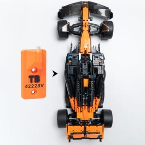 May include: A top-down view of a black and orange Formula 1 race car model next to an orange rectangular plastic tool. The tool features the text TB and 42228V printed in black, with a small peg at the top and two circular holes below.