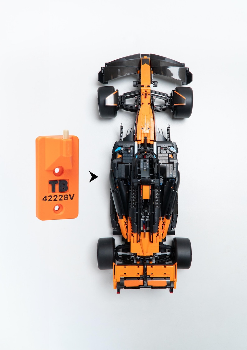 May include: A top-down view of a black and orange Formula 1 race car model next to an orange rectangular plastic tool. The tool features the text TB and 42228V printed in black, with a small peg at the top and two circular holes below.