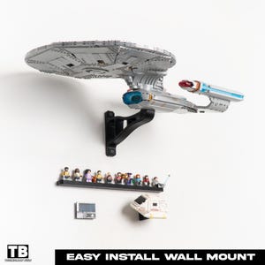 Op de afbeelding: Een zilver en grijs model van het Star Trek USS Enterprise NCC-1701-D ruimteschip gemonteerd op een zwarte muurbeugel. Daaronder toont een plank miniatuurfiguren. De tekst "EASY INSTALL WALL MOUNT" staat onderaan.