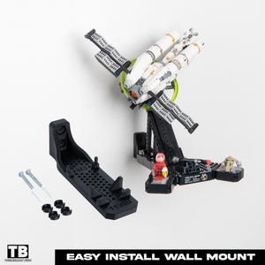 May include: A black wall mount kit for a model spaceship, with a white and gold spaceship model attached. The kit includes a black base, screws, and a small red figurine. The text "EASY INSTALL WALL MOUNT" is visible.