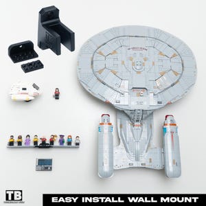 Puede incluir: Maqueta gris y blanca de la USS Enterprise NCC-1701-D de Star Trek, construida con bloques de construcción, con figuras en miniatura y una lanzadera. Incluye componentes de montaje en pared negros. El texto "EASY INSTALL WALL MOUNT" es visible.