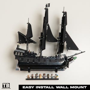 Puede incluir: Modelo de barco pirata negro con velas negras y una bandera de calavera y tibias cruzadas. El barco está montado en una pared con una estantería negra que contiene figuras en miniatura. Se muestra el texto "EASY INSTALL WALL MOUNT".