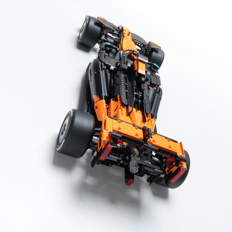 May include: A detailed model race car constructed from orange and black building blocks. The vehicle features a rear spoiler, exposed mechanical components, and four wide black tires. The design includes intricate structural elements and a visible engine assembly at the rear, showcasing a complex engineering build.