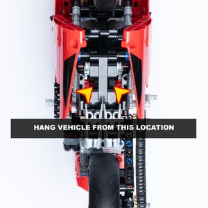 May include: A red and black toy motorcycle with a chain drive and a rear wheel. The motorcycle is hanging from two points on the frame, indicated by yellow arrows. The text "HANG VEHICLE FROM THIS LOCATION" is printed on a black bar below the motorcycle.