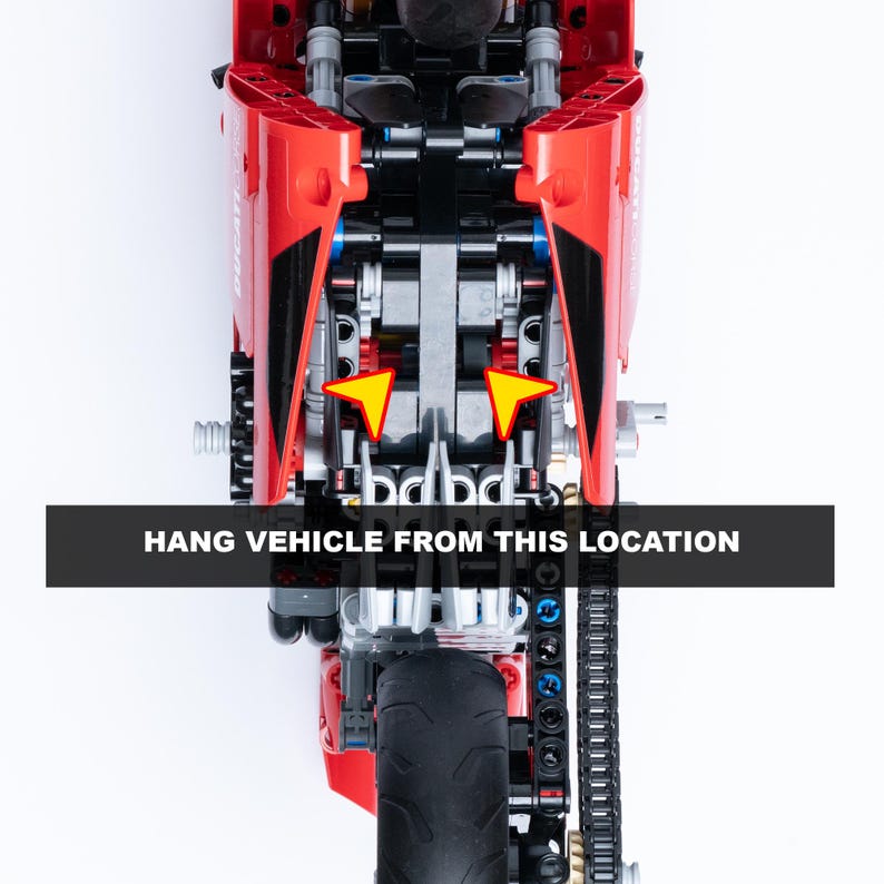 May include: A red and black toy motorcycle with a chain drive and a rear wheel. The motorcycle is hanging from two points on the frame, indicated by yellow arrows. The text "HANG VEHICLE FROM THIS LOCATION" is printed on a black bar below the motorcycle.