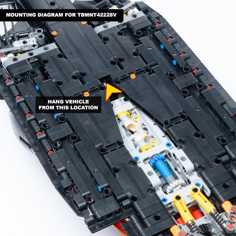 May include: A black plastic construction vehicle model featuring a complex internal gear system with blue, yellow, and white components. Text reads MOUNTING DIAGRAM FOR TBMNT42228V and HANG VEHICLE FROM THIS LOCATION with a yellow arrow pointing to a gap.