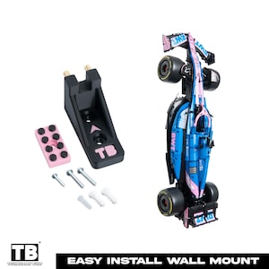 May include: A black plastic wall mount bracket featuring the letters TB, a small pink rectangular component with eight black circular studs, three metal screws, three white plastic wall plugs, and a blue and pink Formula 1 race car model.