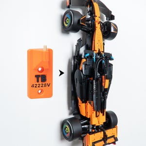 May include: A black and orange Formula 1 race car model with black tires featuring colorful wheel hubs, positioned next to an orange rectangular wall mount bracket labeled with the black text TB 42228V and a small wooden peg attachment.