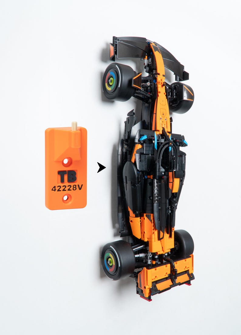 May include: A black and orange Formula 1 race car model with black tires featuring colorful wheel hubs, positioned next to an orange rectangular wall mount bracket labeled with the black text TB 42228V and a small wooden peg attachment.