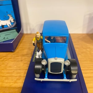 Peut inclure: Un jouet de voiture vintage bleu avec une figurine d'homme en chemise jaune et pantalon marron, et un petit chien blanc. La voiture a des pneus noirs et une calandre avant argentée. Le jouet est sur une surface bleue.