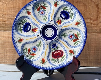 May include: A round ceramic serving plate with six indented sections and a central circular well. The plate features a cream-colored base with hand-painted floral motifs in blue, red, and green, finished with a decorative blue speckled border.