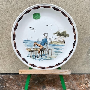 May include: White ceramic plate with a hand-painted seaside scene. The plate features a brown scalloped border and a central image of a person sitting on a pier, with seagulls and sailboats. A green sticker is visible.