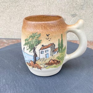 May include: A ceramic mug with a landscape scene. The mug has a tan and white colour scheme with a curved handle. The landscape depicts a house, trees, and a body of water. The mug is likely used for drinking beverages.