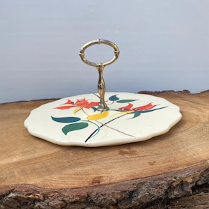 May include: A vintage two-tiered serving tray with a cream-colored surface. The tray features a floral design with red, yellow, and green accents. It has a gold-colored handle. The tray is approximately 8 inches in diameter.