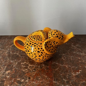 Op de afbeelding: Een keramische theepot met een gele basis en zwarte vlekken, die op een cheetah lijkt. De theepot heeft een gebogen handvat, een tuit en een deksel. De theepot is een decoratief item voor de keuken.