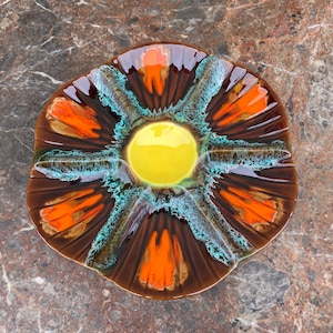 Puede incluir: Un cenicero de cerámica vintage con un diseño único. El cenicero presenta un círculo amarillo central rodeado de acentos marrones, naranjas y turquesas. La forma general es orgánica e irregular, con un acabado brillante. Una pieza decorativa para una estética retro.