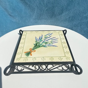 May include: A black metal trivet with a white ceramic tile featuring a bouquet of lavender flowers. The tile has a decorative border of small green leaves.