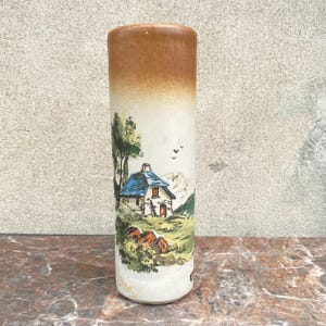 May include: A cylindrical ceramic vase with a landscape scene. The top is brown, fading to white with a painted scene of a house with a blue roof, trees, and mountains. The vase is approximately 8 inches tall.