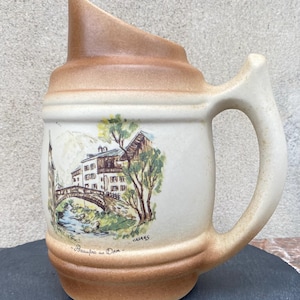 May include: A vintage ceramic beer stein in tan and cream. It has a handle and a slanted spout. The front is decorated with a painted landscape scene of a bridge, buildings, and mountains. The text "Beaufort sur Doron" is also visible on the stein.