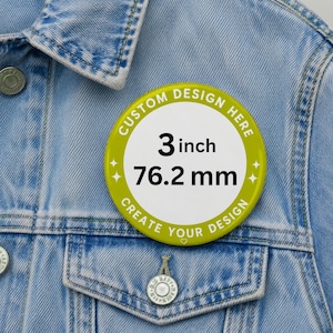 May include: A round pinback button with a lime green border and a white center. The text reads CUSTOM DESIGN HERE, 3 inch, 76.2 mm, and CREATE YOUR DESIGN. The button is attached to a light blue denim jacket with silver metal buttons and stitching.