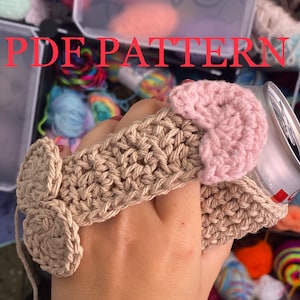 May include: A crocheted beige can cozy with a pink heart on top. The cozy is designed to fit a standard-sized can.
