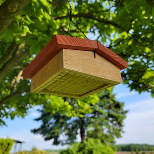 Peut inclure: Un mangeoire à oiseaux en bois avec un toit rouge suspendu à une branche d'arbre. La mangeoire a un fond en grillage métallique et est conçue pour contenir des graines d'oiseaux.