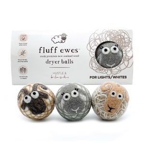 May include: Three wool dryer balls in the shape of sheep with embroidered faces. The balls are beige, gray, and brown. The packaging is white with the text "fluff ewes™ 100% premium new zealand wool dryer balls HUSTLE & blush FOR LIGHTS/WHITES".