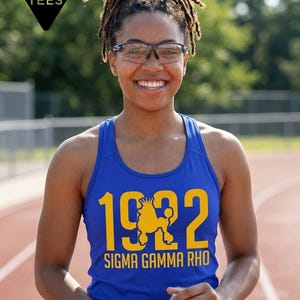 May include: A royal blue racerback tank top featuring the yellow text 1922 with a poodle graphic replacing the zero, and the words SIGMA GAMMA RHO printed below. The top includes a black triangular logo in the corner reading DAY PARTY TEES.