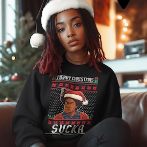 Op de afbeelding: Zwart sweatshirt met een kerstontwerp met de tekst "MERRY CHRISTMAS" en "SUCKA". Het ontwerp bevat een portret van een persoon met een kerstmuts. Het model draagt een zwarte kerstmuts.