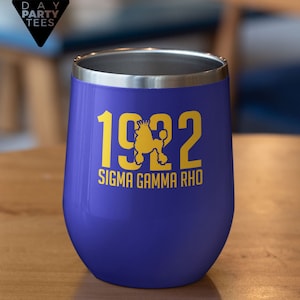 May include: A stemless wine tumbler in a vibrant blue hue, featuring the year "1922" in gold, along with a poodle graphic and the text "SIGMA GAMMA RHO." The tumbler has a silver rim and is likely made of stainless steel.