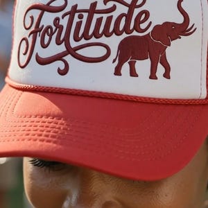 May include: A red and white trucker hat featuring a red embroidered elephant graphic and the word Fortitude in a cursive script font on the front panel. The cap has a solid red brim, a white foam front, and a red mesh back with a red cord trim.
