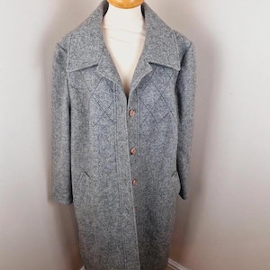 May include: A grey, long coat with a textured finish. The coat has a notched collar, diamond-shaped stitching on the chest, and wooden buttons. It features two front pockets and is displayed on a wooden mannequin.