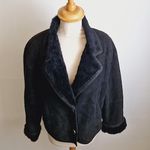 Op de afbeelding: Zwarte suède jas met een kraag en manchetten van imitatiebont. De jas heeft een klassiek ontwerp met een knoopsluiting. De donkere kleur en de pluche afwerking van de jas worden op een mannequin getoond.