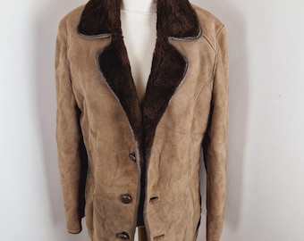 May include: A tan suede jacket with a dark brown faux fur collar and lining. The jacket has a button-up front and is displayed on a wooden mannequin. The jacket appears to be vintage.