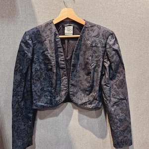 May include: A dark blue cropped jacket with a floral design. The jacket has long sleeves and a V-neck. It is suspended on a wooden hanger. The fabric has a slight sheen. The background is a neutral colour.