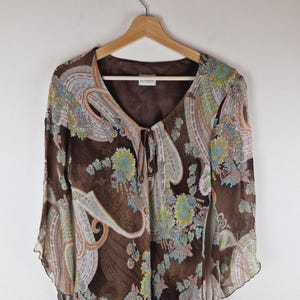 Puede incluir: Una blusa marrón con estampado de cachemira y mangas largas y acampanadas. La blusa presenta un escote en V con cierre de lazo y una tela delicada y fluida. El estampado de cachemira incluye tonos de azul, verde y naranja.